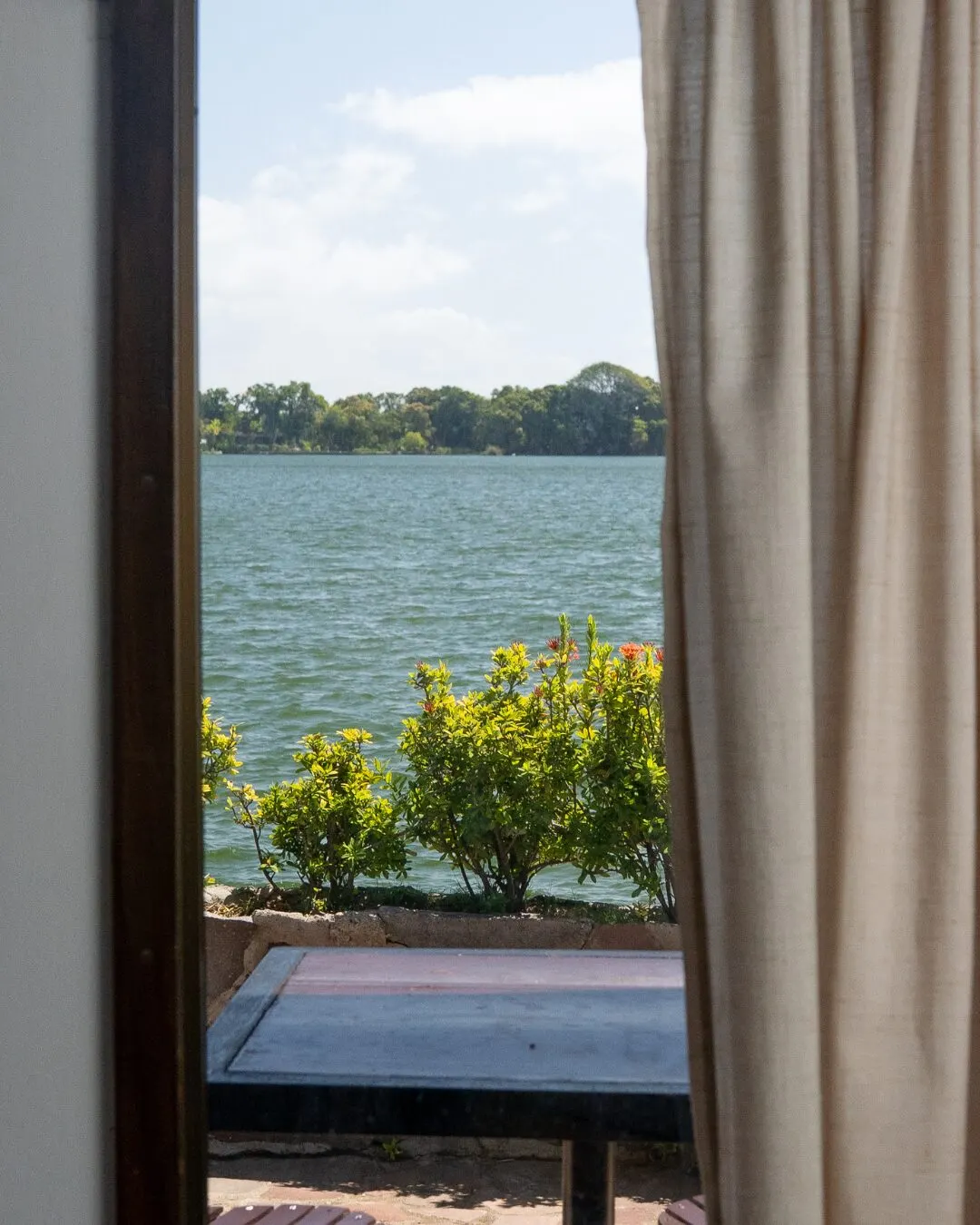 An open window with a curtain, showing the lake and some shrubs.