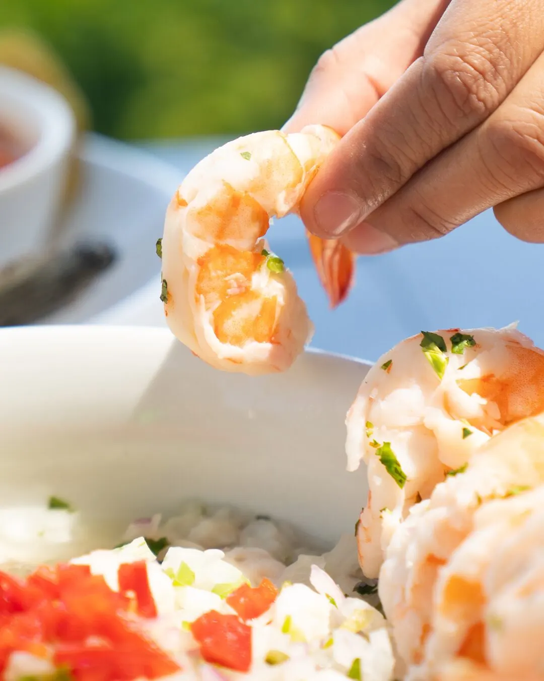 A hand holding a peeled shrimp over a plate of shrimp with pico de gallo.