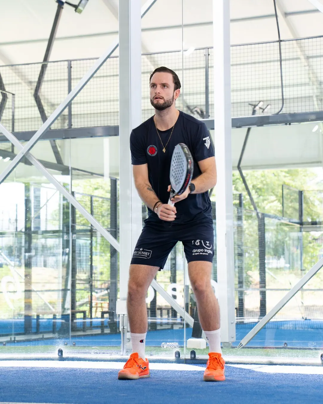 Jugador con camiseta azul marino y tenis naranja en posición de espera, con la pala en la mano, en una cancha azul cubierta.