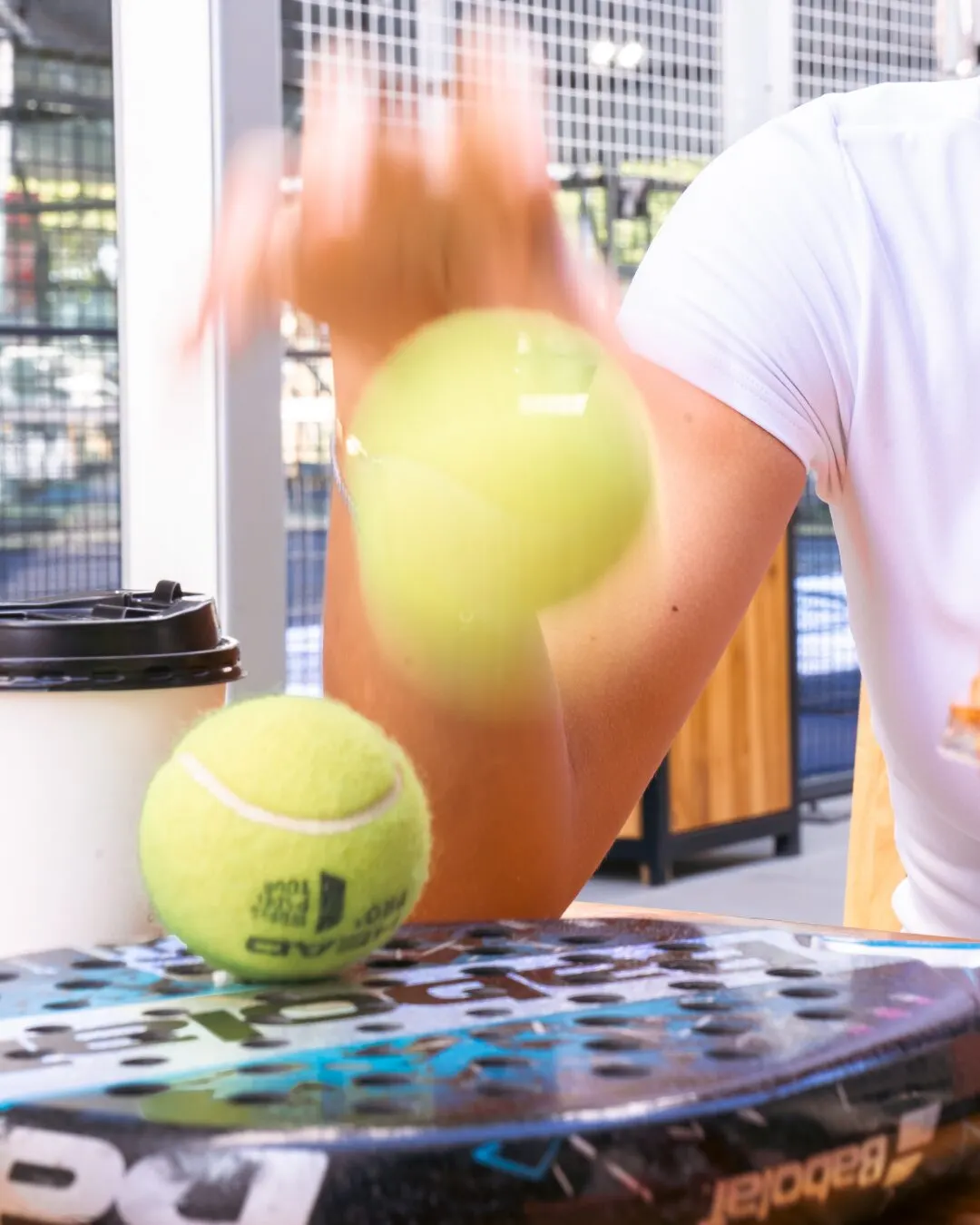 Pelotas amarillas sobre una pala de pádel, con una mano y un vaso de café al fondo.