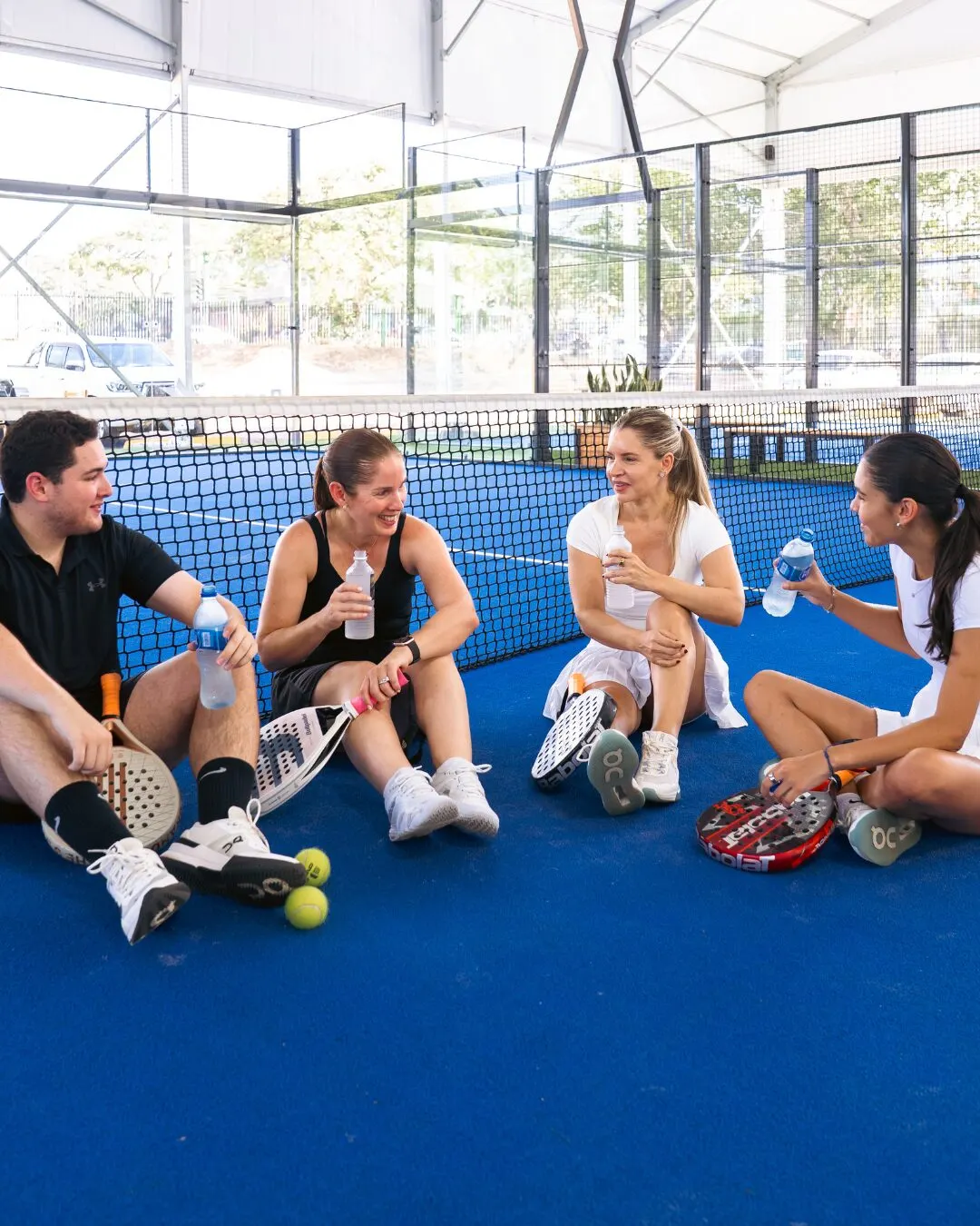 Cuatro personas sentadas en la cancha azul con palas y botellas de agua.