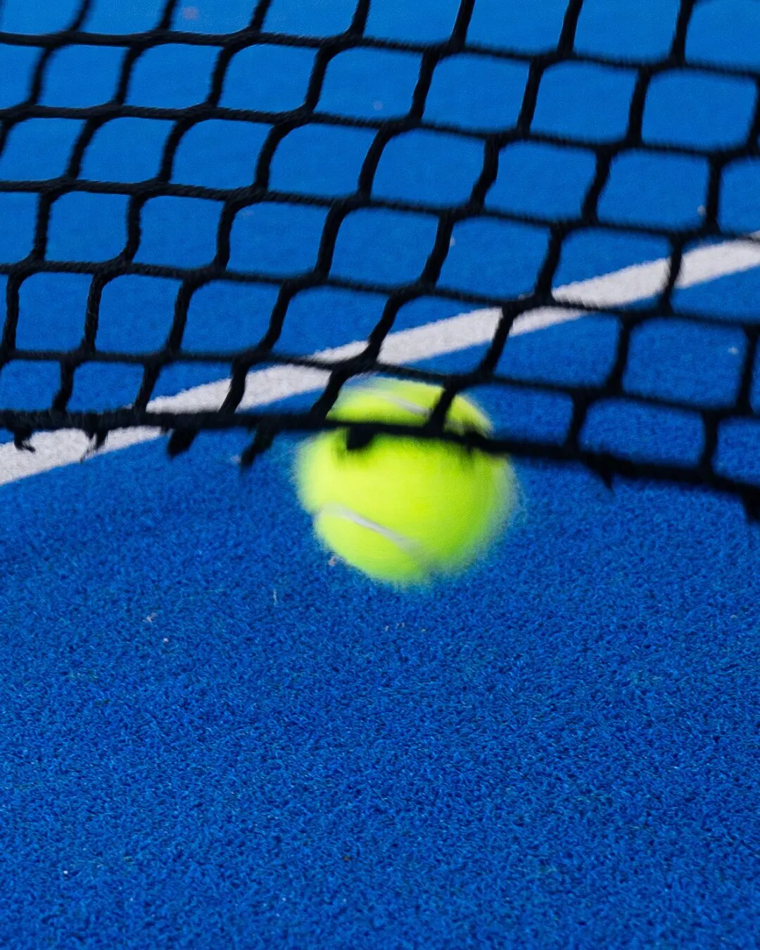 Pelota amarilla pasando sobre la red de una cancha azul.