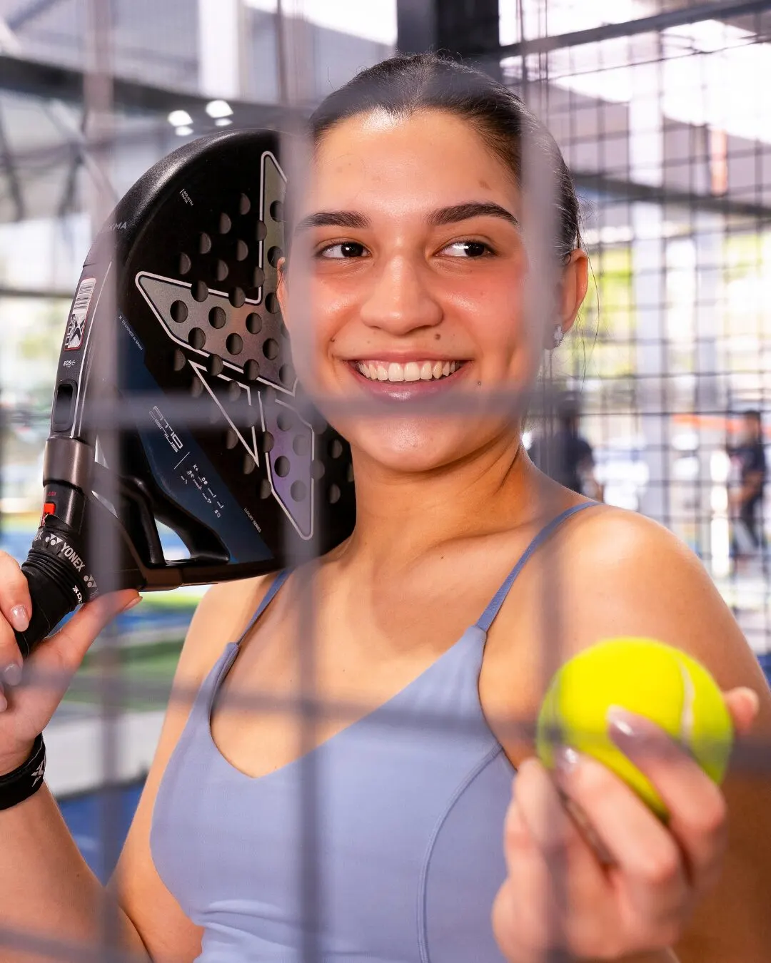 Jugadora sonriente con una pala al hombro y una pelota en la mano, vista a través de la reja.