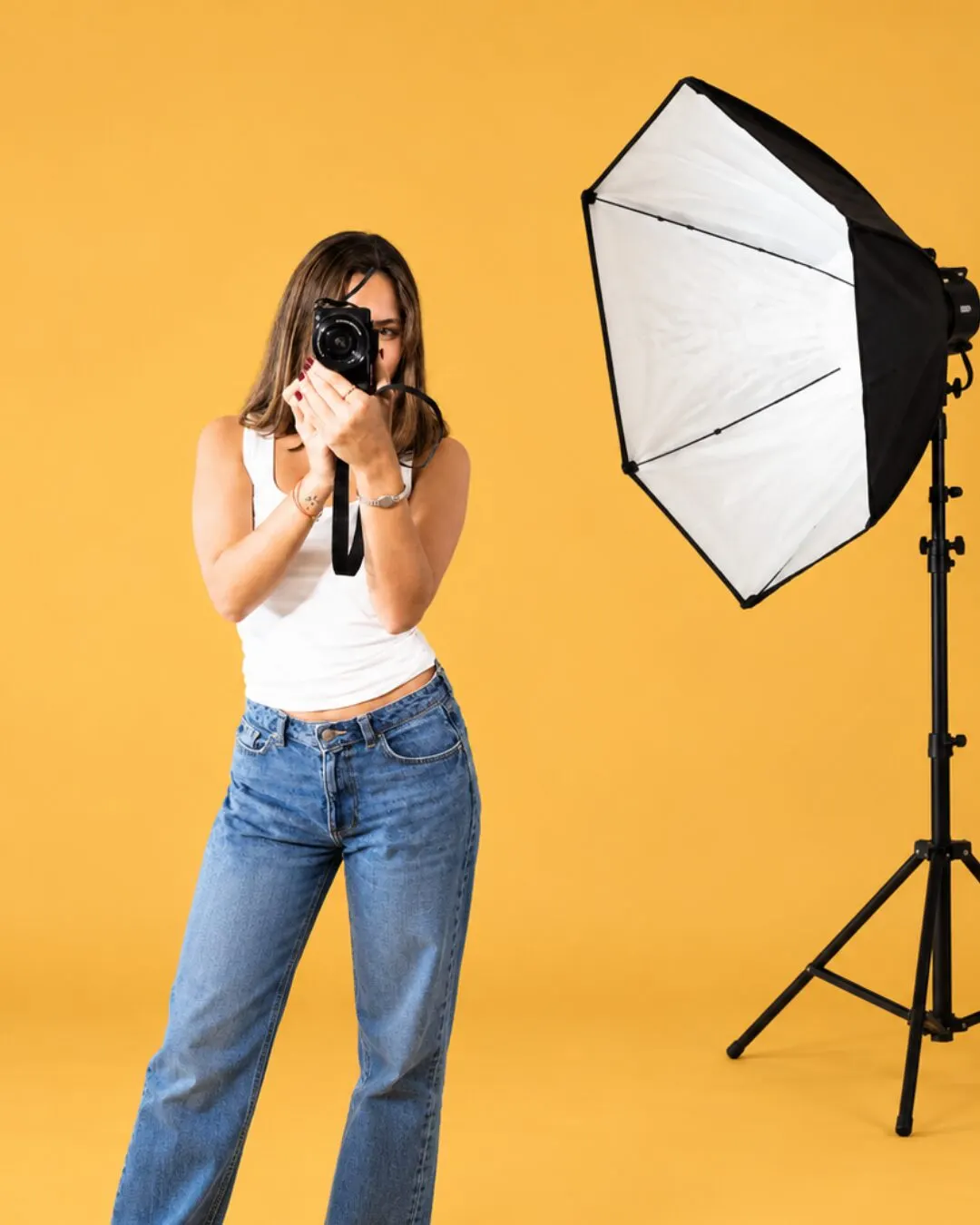 Una fotógrafa apunta su cámara junto a un softbox sobre un fondo mostaza